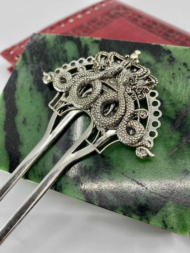 May include: Silver hair fork with a decorative design of a snake with a crown on its head. The fork is on a green and black surface.
