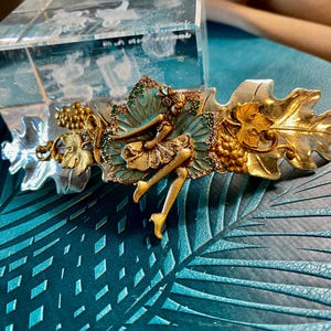 May include: A decorative hair clip featuring a fairy design. The clip has gold, silver, and teal accents, with a fairy figure resting on a leaf. The clip also includes grape clusters and leaf details, creating an elegant accessory.