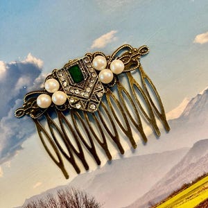 May include: A gold hair comb with a decorative design featuring a green gemstone, rhinestones, and faux pearls.