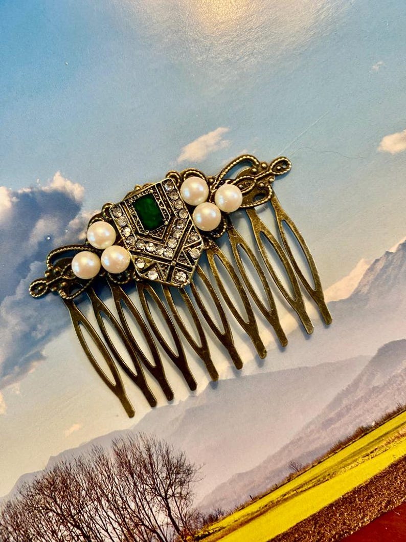 May include: A gold hair comb with a decorative design featuring a green gemstone, rhinestones, and faux pearls.