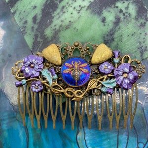 May include: A gold metal hair comb featuring a dragonfly design in the centre. The dragonfly is iridescent blue and purple. The comb is decorated with purple flowers and leaves.