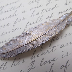 May include: A silver and gold feather hair clip. The feather is detailed with realistic texture and has a subtle sheen.