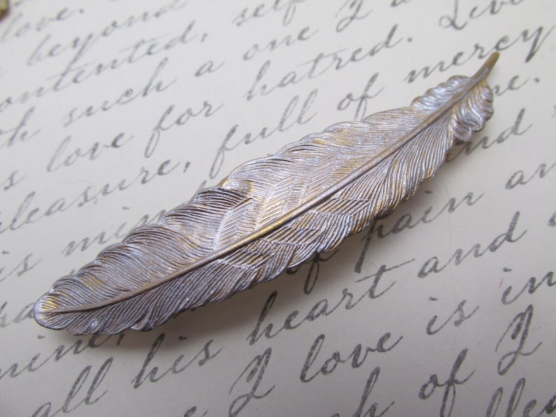 May include: A silver and gold feather hair clip. The feather is detailed with realistic texture and has a subtle sheen.