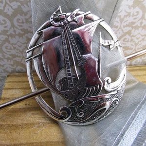 May include: A silver-toned hair stick featuring a detailed ship design within a circular frame. The ship has visible masts, sails, and rigging. The stick is inserted through a gray fabric, suggesting its use as a hair accessory. The background is a light beige pattern.