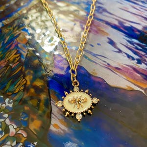 Könnte beinhalten: Goldfarbene Halskette mit einem ovalen Anhänger. Der Anhänger hat eine cremefarbene Mitte mit einem goldenen Bienenmuster, umgeben von klaren Kristallen. Die Kette ist im Chunky-Link-Stil gehalten.