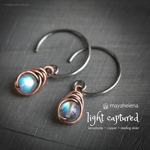 May include: A pair of hoop earrings with a copper wire-wrapped design. Each earring features a blue and green iridescent labradorite stone. The hoops are made of sterling silver. The text "light captured" is visible.
