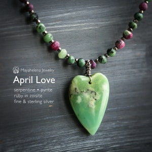 Pode incluir: Um colar com um pingente de coração de serpentina verde, contas de rubi em zoisita e detalhes de pirita. O colar é feito de prata fina e de lei. O pingente tem a forma de um coração e é de cor verde e branca. O colar está sobre um fundo de madeira.  "April Love" está escrito na imagem.