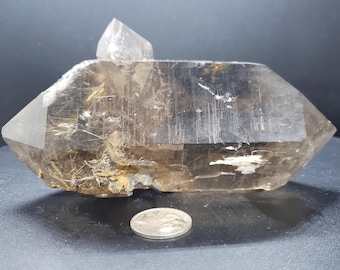 May include: A large, clear quartz crystal with a double termination and inclusions of golden rutile. The crystal is approximately 4 inches long and is sitting on a dark surface next to a US quarter for scale.