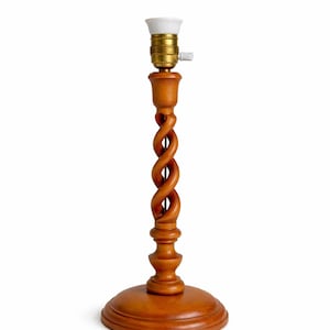 Könnte beinhalten: Eine Tischlampe aus Holz mit einer Lampenfassung. Die Lampe hat einen runden Sockel und einen gedrehten, geschnitzten Stiel. Die Lampe ist aus hellbraunem Holz gefertigt und hat eine weiße und goldfarbene Fassung.