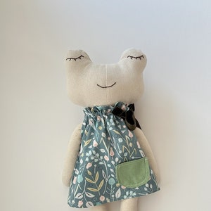 Peut inclure: Poupée grenouille faite à la main avec une tête et des membres beiges, et une robe turquoise à motifs floraux. La poupée a les yeux fermés et un sourire cousu. Une poche verte est cousue sur la robe.