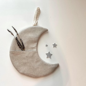May include: A beige crescent moon-shaped wall hanging with a pocket, holding dried lavender stems. Three silver glitter star decorations are next to the moon. A cream-coloured lace ribbon is attached for hanging.