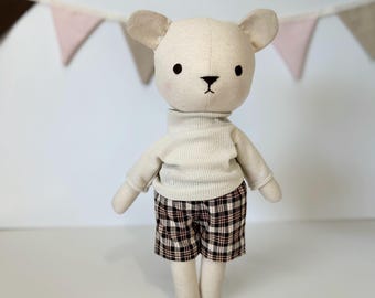 May include: A plush white bear wearing a beige turtleneck sweater and plaid shorts. The bear has black eyes and a black nose.