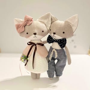 May include: Two handmade fox dolls. One doll wears a pink bow, a pink cape, and a white dress with a floral bouquet. The other doll wears a black and white polka dot bow tie and gray pants. Both dolls are holding hands.