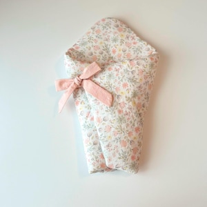 May include: A floral print baby swaddle blanket with a pink bow. The blanket features a pattern of small pink, yellow, and green flowers on a white background. The bow is a solid light pink color.