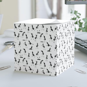 May include: A white square notepad covered in a repeating pattern of black cartoon ants in various poses. The ants are stylized with simple lines and shapes, creating a playful and whimsical design. The notepad is sitting on a white surface.