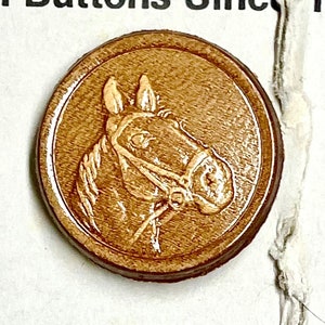 May include: Two leather buttons with a horse head design. The top button is brown with a light brown horse head. The bottom button is red with a dark red horse head.