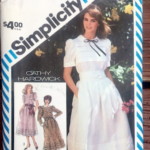 May include: Simplicity sewing pattern number 5974 for a dress with a white background and blue and white striped border. The pattern is for a Cathy Hardwick design and costs $4.00. The pattern shows a white dress with a black bow and a white collar. The dress has a gathered skirt and a fitted bodice.