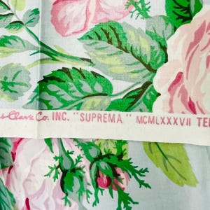 May include: Floral fabric featuring large pink roses with green leaves on a light blue background. A white strip across the fabric has text that reads "Cyrus Clark Co. INC. SUPREMA" and other text.