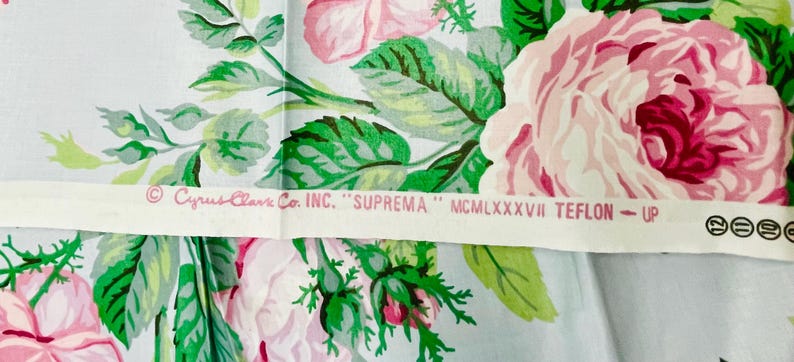 May include: Floral fabric featuring large pink roses with green leaves on a light blue background. A white strip across the fabric has text that reads "Cyrus Clark Co. INC. SUPREMA" and other text.