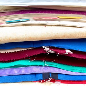 Pode incluir: Uma pilha de amostras de tecido e papéis em várias cores. Os tecidos incluem azul, bordô, verde-azulado e roxo. Papéis com abas são visíveis no topo. A imagem sugere uma coleção de têxteis ou amostras de design.