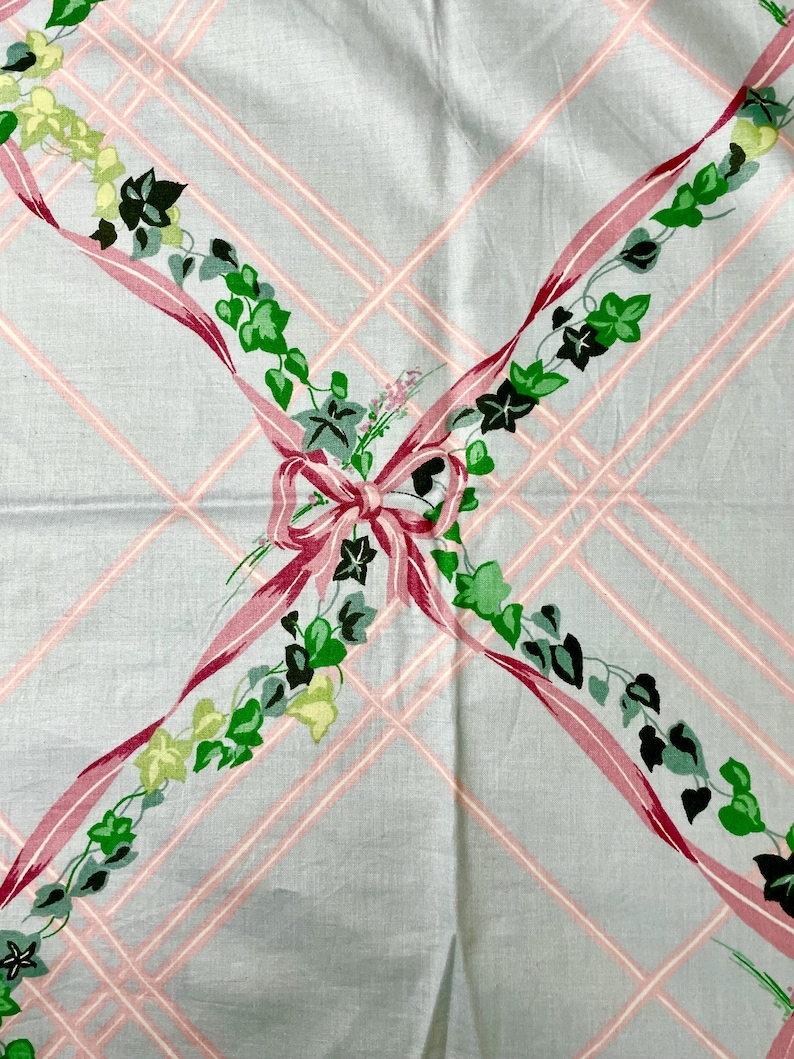 May include: A vintage light blue tablecloth with a pink plaid pattern. Pink ribbons with green ivy and a pink bow decorate the tablecloth. The tablecloth is made of a soft, possibly cotton, material.
