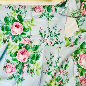 May include: Light blue fabric with a floral pattern featuring pink roses and green leaves. The fabric is cut into various pieces, suggesting it's for sewing or crafting projects. The design is a classic floral print.