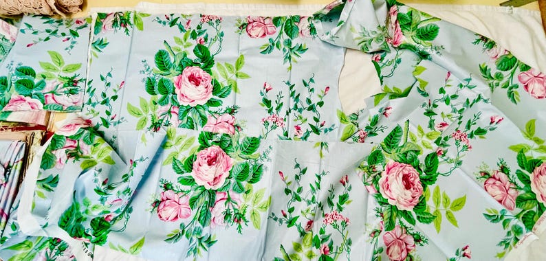 May include: Light blue fabric with a floral pattern featuring pink roses and green leaves. The fabric is cut into various pieces, suggesting it's for sewing or crafting projects. The design is a classic floral print.