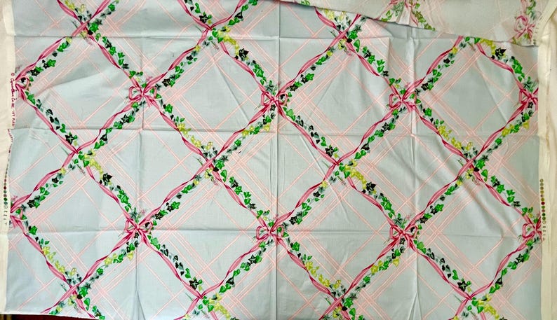 May include: Light blue fabric with a repeating pattern of pink ribbons, green vines, and bows. The design is arranged in a grid pattern with pink lines. The fabric appears to be cotton or a similar material.