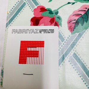 May include: A floral patterned handkerchief with pink roses and green leaves. The background has a light green and blue geometric design. A white card with the text "FABRIYAZ" and address information is in the foreground.