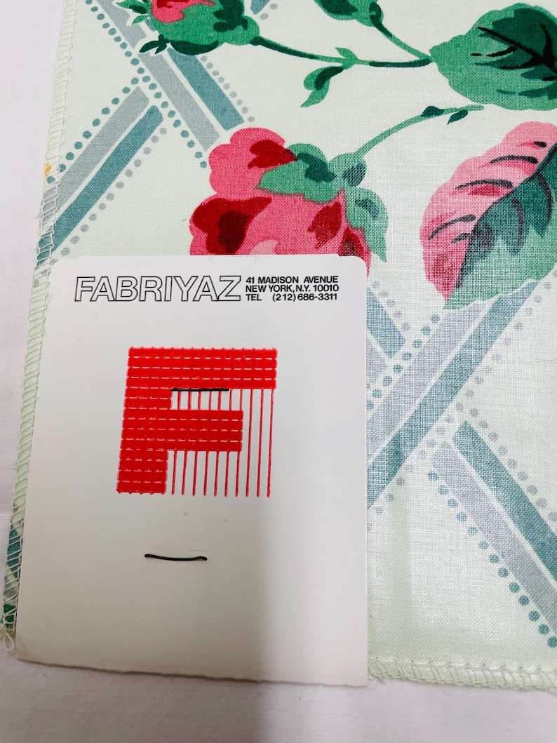 May include: A floral patterned handkerchief with pink roses and green leaves. The background has a light green and blue geometric design. A white card with the text "FABRIYAZ" and address information is in the foreground.