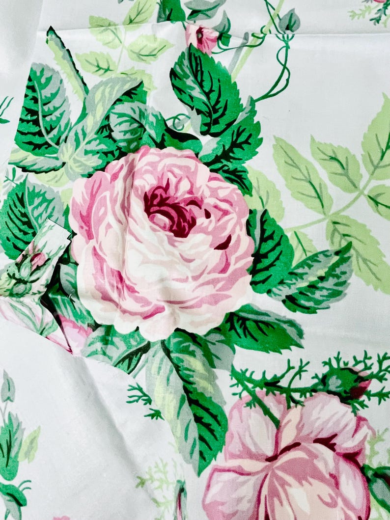 May include: White fabric with a repeating floral pattern. Large pink roses with dark pink centers are surrounded by green leaves and vines. The design has a vintage, botanical feel.