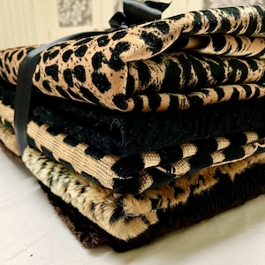 May include: A stack of folded fabric with various animal print patterns, including leopard and zebra designs, in shades of tan, black, and brown. A black ribbon wraps around the stack, securing the fabrics. The fabrics appear to be soft and textured.