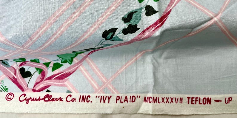 May include: Light blue fabric with a pink plaid pattern and floral accents. The fabric features a design of ivy leaves and pink ribbons. The text "Cyrus Clark Co. INC. 'IVY PLAID'" is printed at the bottom.