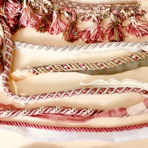 May include: An assortment of decorative trims in various styles and colors. The image showcases several types of trim, including fringed, braided, and tasseled designs. Colors range from deep reds and pinks to neutral tones, suitable for craft and sewing projects.