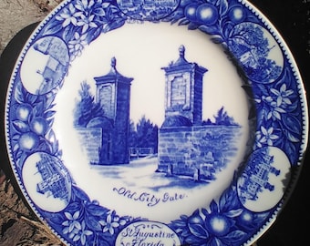 May include: A white and blue decorative plate featuring the "Old City Gate" in St. Augustine, Florida. The plate has a blue border with floral and fruit designs, and includes text that reads "St. Augustine Florida Settled 1565."