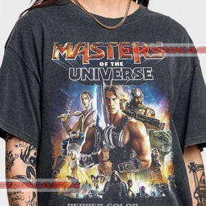 Könnte beinhalten: Dunkelgraues T-Shirt mit einem farbenfrohen Grafikdesign von "Masters of the Universe". Das Design zeigt Charaktere mit Schwertern und ein löwenartiges Wesen, mit dem Text "MASTERS OF THE UNIVERSE". Der Text "PEPPER COLOR" befindet sich unten.