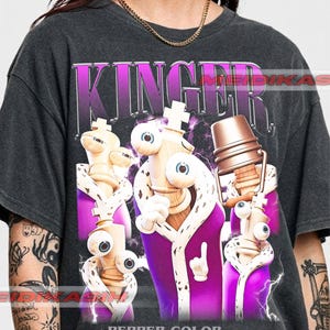 May include: A charcoal gray t-shirt featuring a graphic print of Kinger from The Amazing Digital Circus. The design displays the character in multiple poses wearing a purple robe with white trim, surrounded by purple lightning and the text KINGER.