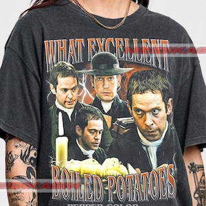Może przedstawiać: Ciemnoszary t-shirt z krótkim rękawem z nadrukiem przedstawiającym czterech mężczyzn w strojach z epoki oraz napisem "WHAT EXCELLENT BOILED POTATOES" i "PEPPER COLOR". Wzór zawiera zapaloną świecę oraz miskę ziemniaków na pierwszym planie.