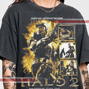 Könnte beinhalten: Ein anthrazitfarbenes T-Shirt mit einer gelb-schwarzen Grafik eines futuristischen gepanzerten Soldaten. Der Text lautet "EARTH WILL NEVER BE THE SAME" und "HALO 2". Drei kleine rechteckige Felder zeigen Actionszenen, und "PEPPER COLOR" ist unten aufgedruckt.