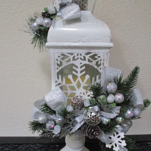 Puede incluir: Linterna blanca con un diseño de copo de nieve y un lazo plateado. La linterna está decorada con una cinta plateada, ramas de pino y copos de nieve blancos. La linterna está sentada sobre una base blanca.