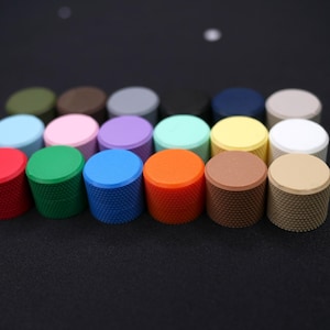 Puede incluir: Una colección de perillas cilíndricas coloridas con lados texturizados y tapas planas. Las perillas son de varios colores, incluyendo rojo, verde, azul, naranja, marrón y beige. Están dispuestas en filas sobre una superficie oscura.