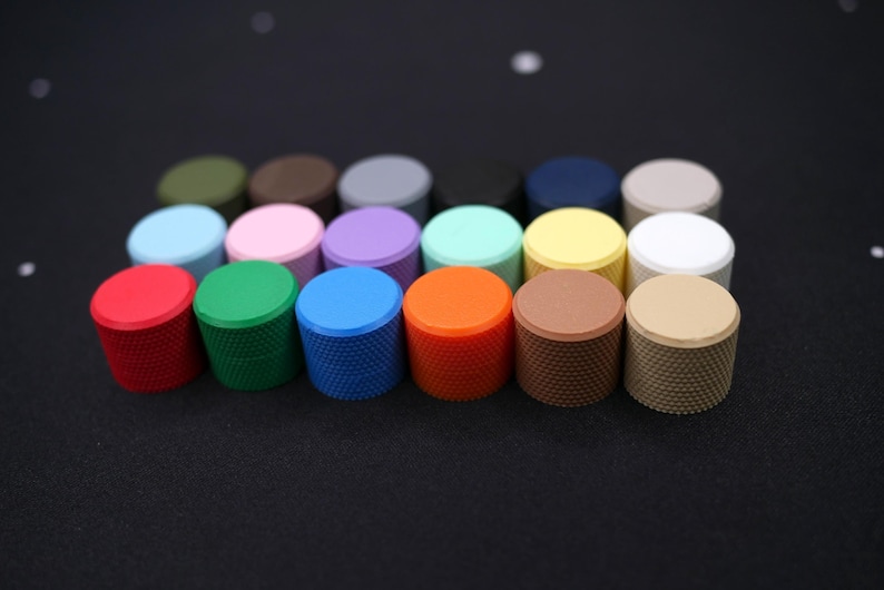 May include: A collection of colorful, cylindrical knobs with textured sides and flat tops. The knobs are in various colors, including red, green, blue, orange, brown, and beige. They are arranged in rows on a dark surface.