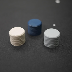 May include: Three cylindrical knobs with textured sides and flat tops are arranged on a dark surface. One is off-white, one is navy blue, and the third is light gray. The knobs appear to be made of metal.