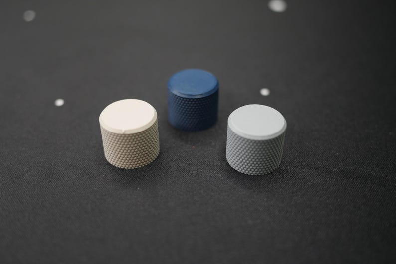 May include: Three cylindrical knobs with textured sides and flat tops are arranged on a dark surface. One is off-white, one is navy blue, and the third is light gray. The knobs appear to be made of metal.