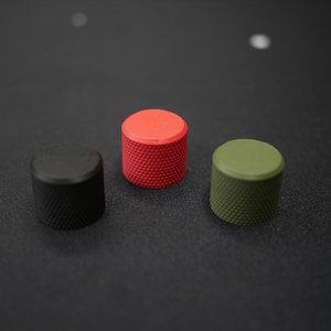 May include: Three cylindrical knobs in a row, each with a textured surface. The knobs are black, red, and olive green. The knobs are on a dark gray surface.