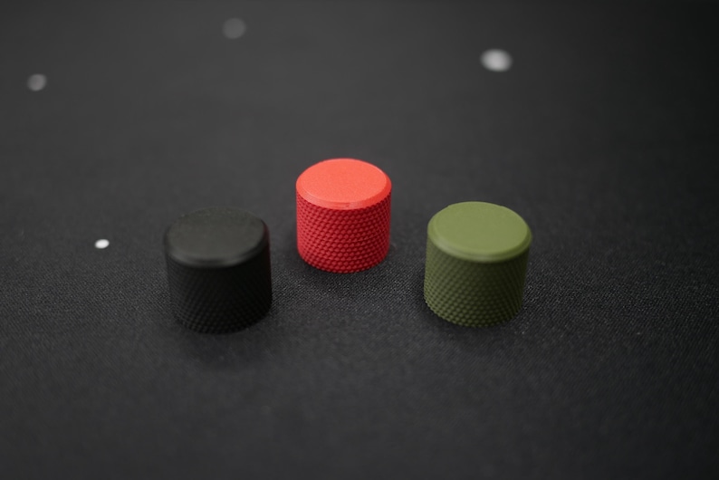 May include: Three cylindrical knobs in a row, each with a textured surface. The knobs are black, red, and olive green. The knobs are on a dark gray surface.