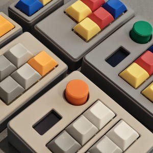 May include: Close-up of several custom mechanical keyboard keypads. Each keypad features a variety of colored keycaps, including blue, red, yellow, orange, and gray. Some keypads have a single round button in orange or green.
