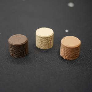 May include: Three cylindrical knobs in shades of brown and beige are arranged on a dark surface. Each knob has a textured, dotted pattern on its sides and a smooth, flat top. The knobs vary in color from dark brown to light beige.