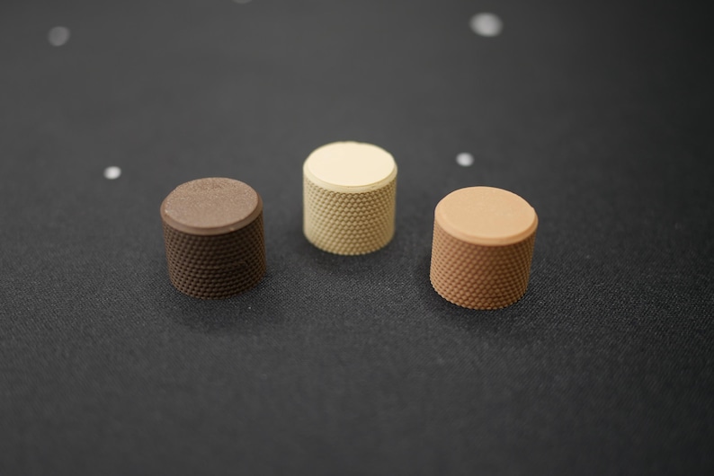 May include: Three cylindrical knobs in shades of brown and beige are arranged on a dark surface. Each knob has a textured, dotted pattern on its sides and a smooth, flat top. The knobs vary in color from dark brown to light beige.