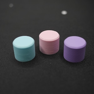 May include: Three colorful knobs in a row. The knobs are cylindrical with a textured surface. One is light blue, one is pink, and one is purple. The knobs are on a dark surface.
